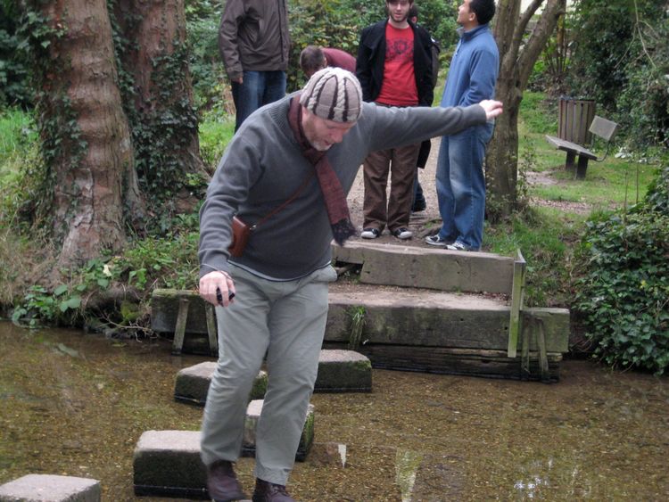 Man clad in outdoor-gear, walking on stepping-stones across shallow water; others lined up to follow