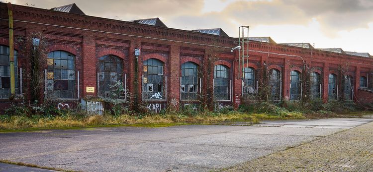 Red brick industrial building, arched, paned windows, on an asphalt lot, cloudy sky, weeds and graffitti