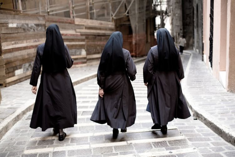 Three nuns, in traditional Catholic habits, viewed from behind, walking on a narrow cobblestone street.