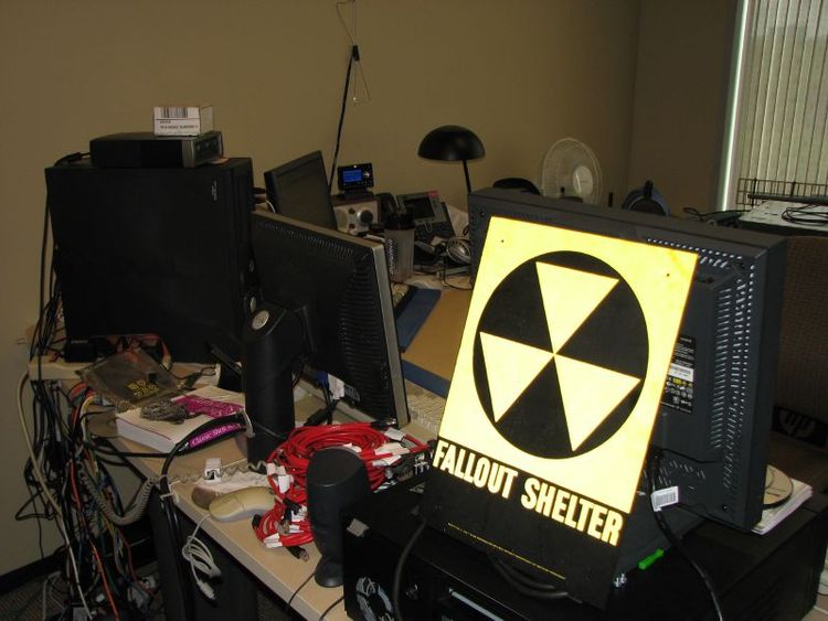 Messy office! The backside of multiple computer terminals with piles of wire, yellow/black "Fallout Shelter" sign.