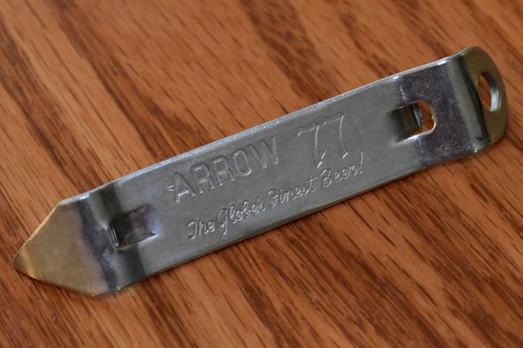 Old-fashioned silver-colored can opener, stamped with "ARROW 77, The Globe's Finest Beer!" on a woodgrain background.