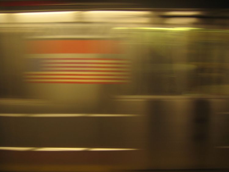 A NYC subway train streaks by, blurry, metal; the exposure elongates the US flag emblem on the side of the train