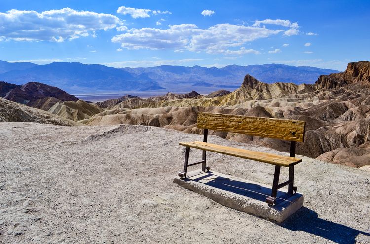 Craggy wooden bench on dusty sand, with desert mountains in the background.  Sunny day, blue sky, low clouds