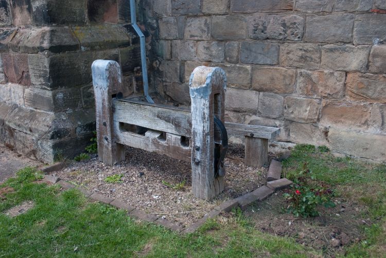 Medieval stocks, in front of an English church. Old wood, on gravel, bricks of varying rust and grey, green grass and plants.