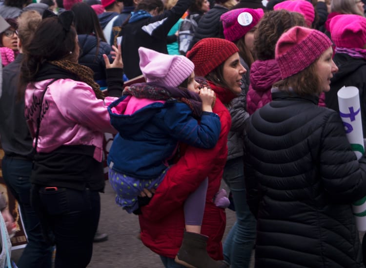 Women in a crowd wearing lots of pink, pink-hatted young girl piggyback with her mom, in red.