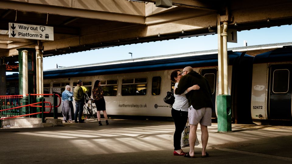 A couple embraces at a UK railway station, train in the background, light filtering through skylight.