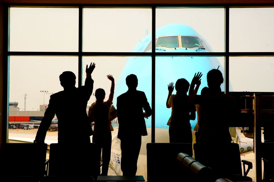 Silhouettes of 5 people waving, backdrop is an airport window at the gate, a huge blue plane in background