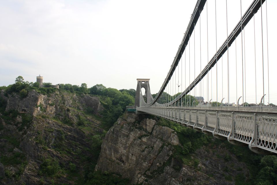 Suspension bridge spanning rocky landscape, with some greenery, white-blue sky, tower-like structure on the horizon