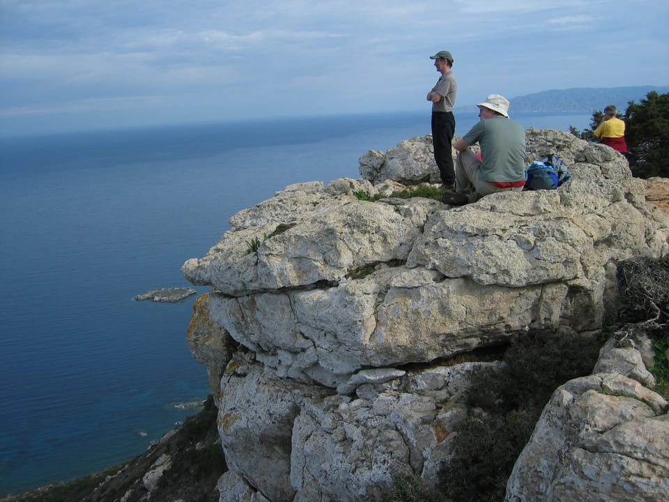 Two men in foreground on craggy limestone (?) cliffs, looking out at blue sky, blue sea. Woman in background.