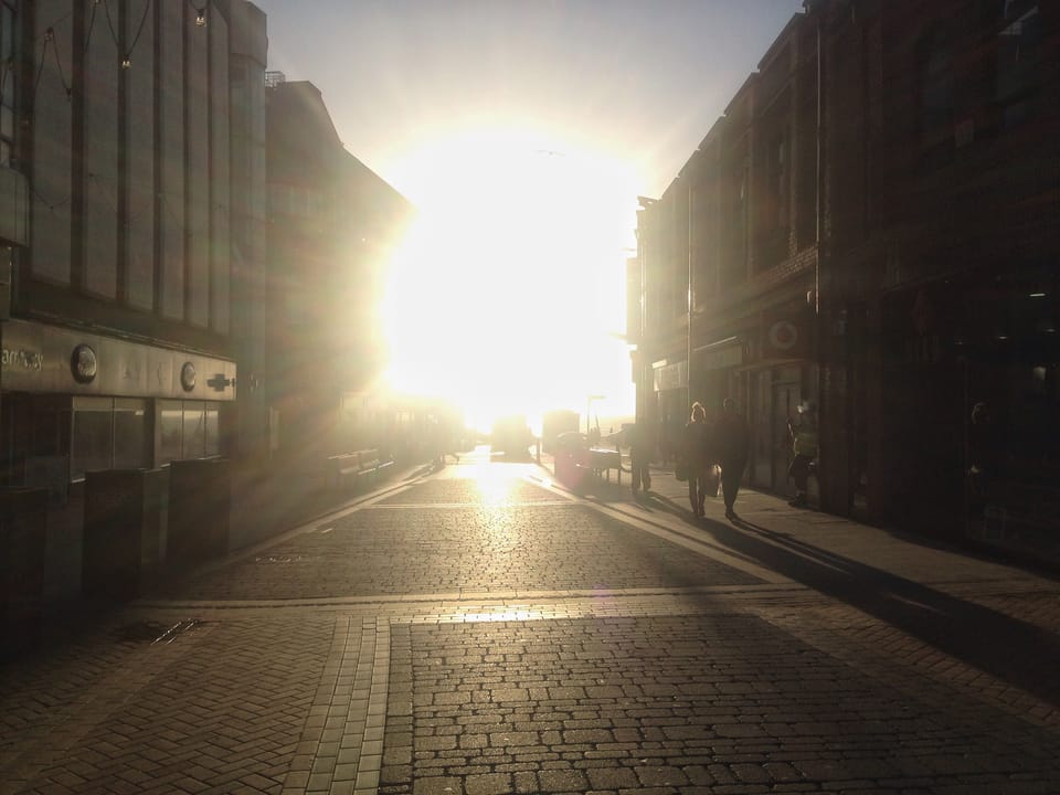 Bright sun corona, coming straight down a cobblestone street, grey-browh buildings and street framing the sun(rise?)